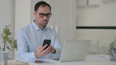 Bilgisayarda çalışırken akıllı telefon kullanan orta yaşlı bir adam