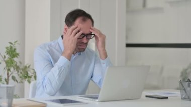 Başağrısı olan orta yaşlı bir adam ofiste dizüstü bilgisayarla çalışıyor.