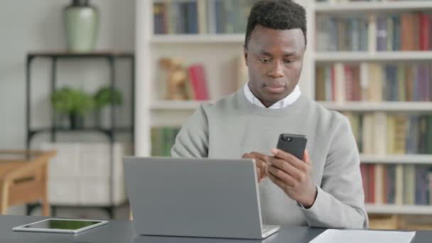 Afričan používající smartphone při použití notebooku v knihovně — Stock video
