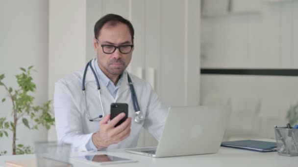 中老年医生在手提电脑上一边用智能手机一边说话 — 图库视频影像