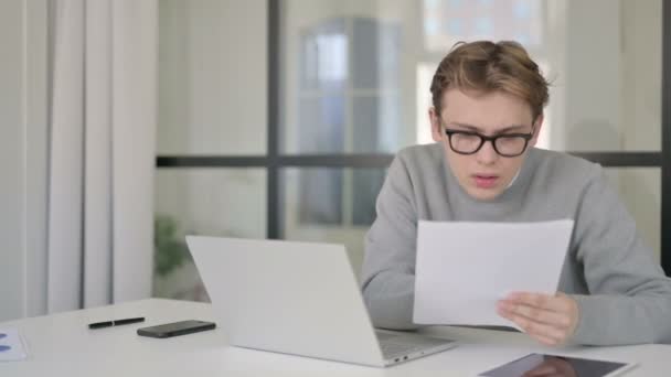 Fiatal férfi laptoppal reagál a veszteségre, miközben dokumentumokat olvas az irodában — Stock videók