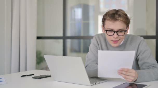 オフィスで文書を読んでいる間、ラップトップの成功を祝う若い男 — ストック動画