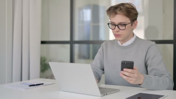 Молодой человек с помощью смартфона при использовании ноутбука в современном офисе — стоковое видео