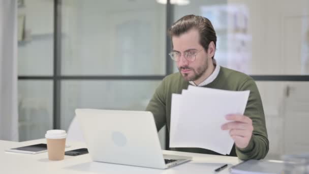 Jeune homme d'affaires avec ordinateur portable lecture de documents dans le bureau — Video