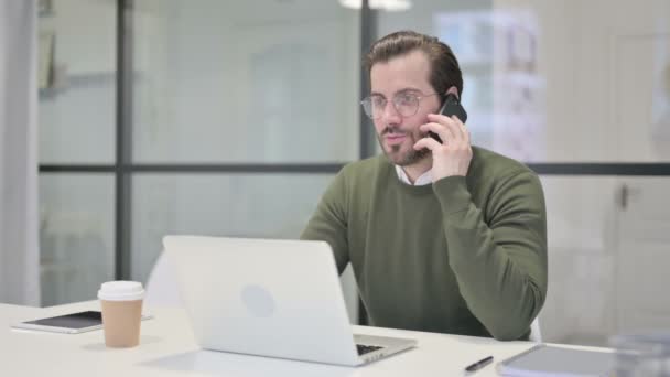 Jeune homme d'affaires parlant sur Smartphone tout en utilisant un ordinateur portable dans le bureau — Video