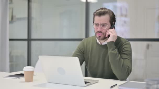 Wütender junger Geschäftsmann spricht auf Smartphone, während er Laptop im Büro benutzt — Stockvideo