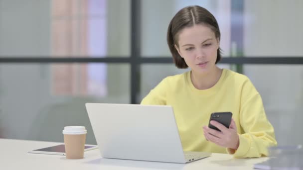 Mujer joven usando Smartphone mientras usa el ordenador portátil en la oficina — Vídeo de stock