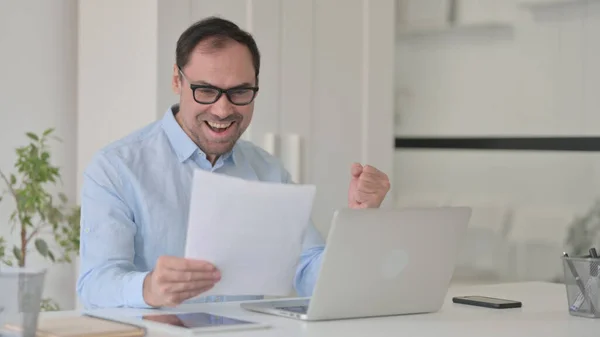 Erfolgreicher Mann mittleren Alters liest Dokumente mit Laptop — Stockfoto