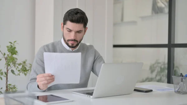 Junger Mann mit Laptop liest Dokumente im Büro — Stockfoto