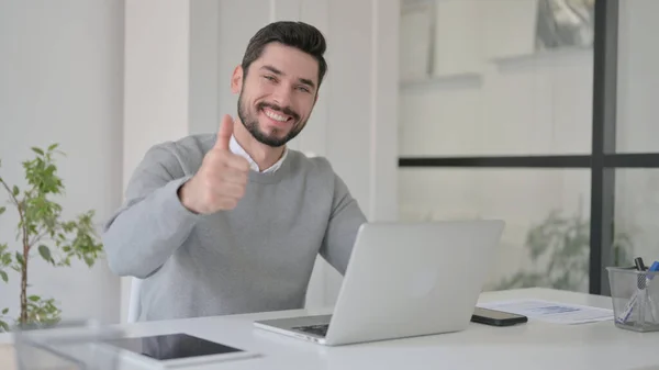 Hombre joven mostrando pulgares hacia arriba firmar mientras se utiliza el ordenador portátil en el trabajo — Foto de Stock