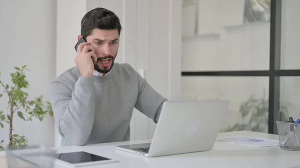 Wütender Geschäftsmann spricht auf Smartphone, während er Laptop im Büro benutzt — Stockfoto