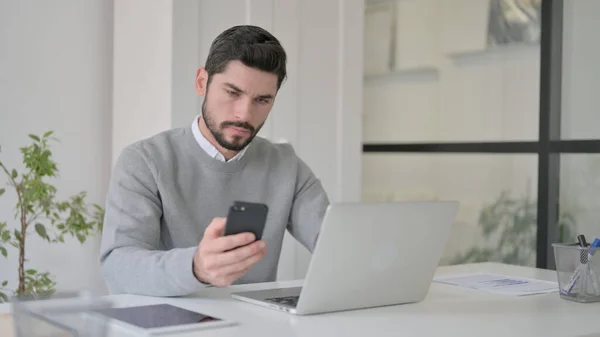 Fiatal férfi használja Smartphone használata közben Laptop az irodában — Stock Fotó