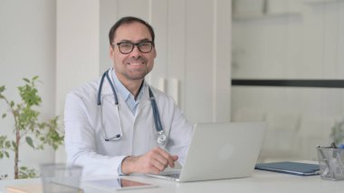 Bilgisayarlı orta yaşlı doktor klinikteki kameraya gülümsüyor.