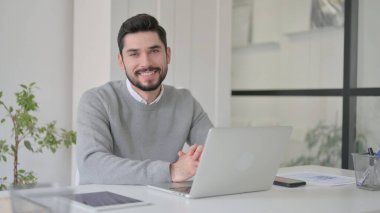 Ofiste dizüstü bilgisayar kullanırken kameraya gülümseyen genç adam