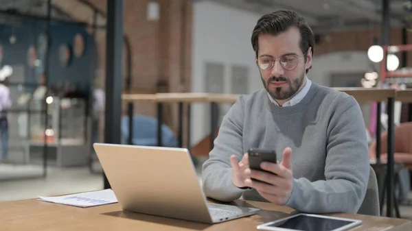 Молодой человек с помощью смартфона и ноутбука в офисе — стоковое фото