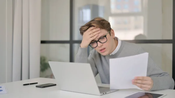Młody człowiek z laptopem reagujący na utratę podczas czytania dokumentów w biurze — Zdjęcie stockowe