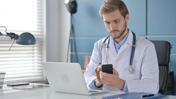 Médico usando Smartphone mientras trabaja en el ordenador portátil en la clínica —  Fotos de Stock
