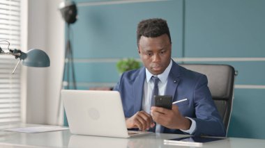 Ofiste dizüstü bilgisayar kullanırken akıllı telefon kullanan Afrikalı iş adamı 