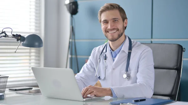 Доктор с ноутбуком улыбается перед камерой в клинике — стоковое фото