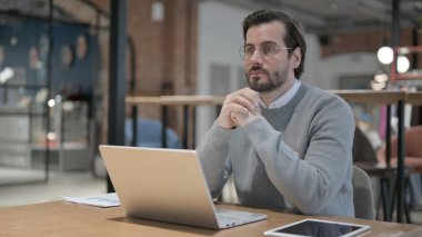 Ofiste dizüstü bilgisayarla çalışırken düşünen genç adam.