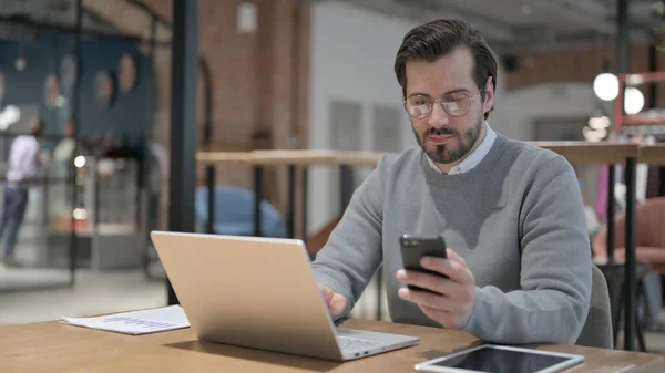 Молодой человек с помощью смартфона во время использования ноутбука в офисе — стоковое фото