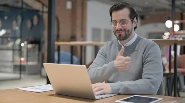 Hombre joven mostrando pulgares hacia arriba firmar mientras se utiliza el ordenador portátil en el trabajo — Foto de Stock