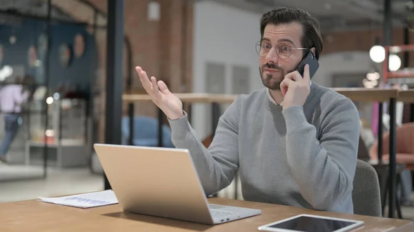 Hombre joven hablando por teléfono mientras usa el ordenador portátil en la oficina — Foto de Stock
