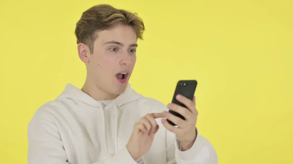 Young Man Surprised by Results on Smartphone on Yellow Background — Photo