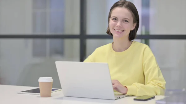 Jovem mulher sorrindo para a câmera enquanto usa laptop no escritório — Fotografia de Stock