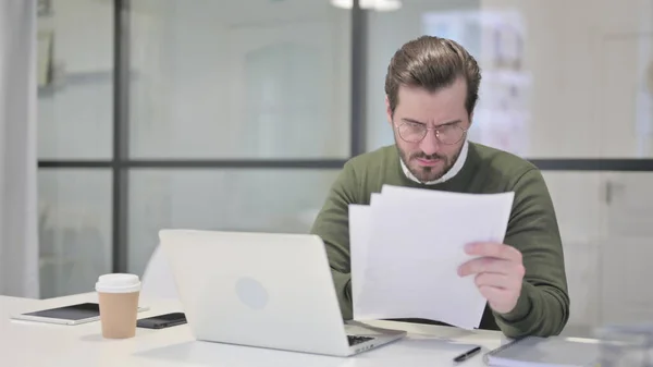 Junger Geschäftsmann mit Laptop liest Dokumente im Büro — Stockfoto