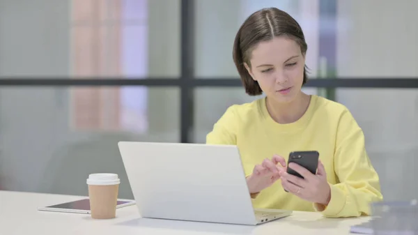 Młoda kobieta korzystająca ze smartfona podczas korzystania z laptopa w biurze — Zdjęcie stockowe