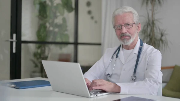 Senior Old Doctor Looking at Camera while using Laptop in Clinic — Fotografia de Stock