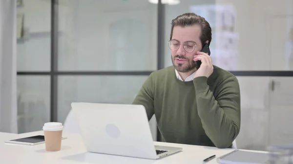 Giovane uomo d'affari che parla su smartphone durante l'utilizzo del computer portatile in ufficio — Foto Stock