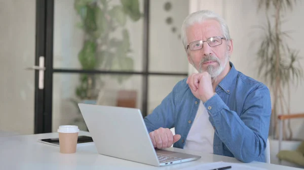 Anciano mayor mirando a la cámara mientras usa el ordenador portátil en la oficina —  Fotos de Stock