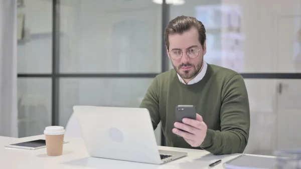 Jonge zakenman met behulp van Smartphone tijdens het gebruik van Laptop in Office — Stockfoto