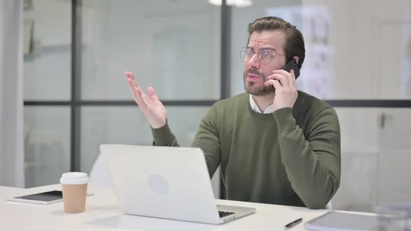 Boos jonge zakenman praten op Smartphone tijdens het gebruik van Laptop in Office — Stockfoto