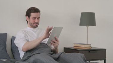 Genç adam yatakta otururken tablet kullanıyor.