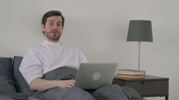 Fiatal férfi laptoppal Nézi a kamerát az ágyban — Stock videók