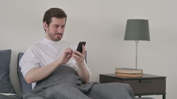 ベッドでスマートフォンで成功を祝う若い男 — ストック動画