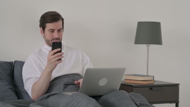 ベッドの中でスマートフォンを使用してノートパソコンを持つ若い男 — ストック動画