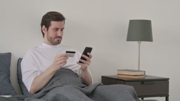Молодой человек совершает онлайн-оплату на смартфоне в постели — стоковое видео