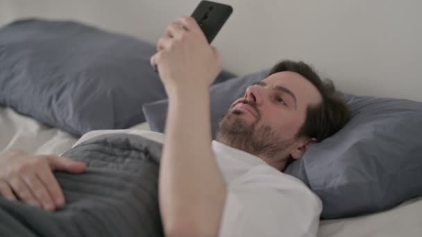 Młody człowiek za pomocą smartfona podczas snu w łóżku — Wideo stockowe