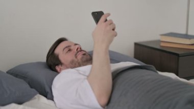 Genç adam yatakta yatarken akıllı telefon kullanıyor.