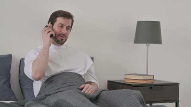 若い男はベッドの中でスマートフォンで通話中 — ストック動画