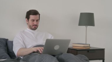 Dizüstü bilgisayarı olan genç adam yatakta baş ağrısı çekiyor.