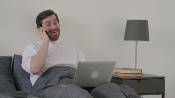 ベッドの中でスマートフォンで話すラップトップを持つ若い男 — ストック動画