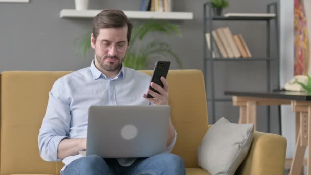 Jovem com laptop usando Smartphone no sofá — Vídeo de Stock