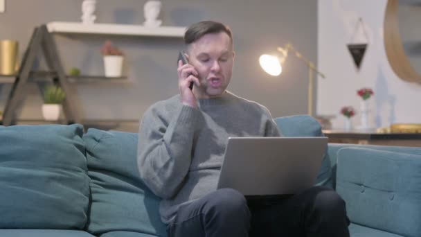 Angry Casual Manwith Laptop Talking on Smartphone на сайті Sofa — стокове відео