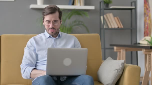 Hombre joven con ordenador portátil reaccionando a la pérdida en el sofá — Vídeos de Stock