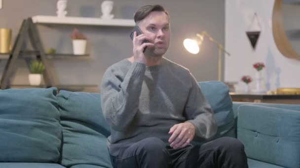 怒っているカジュアル男はソファの上でスマートフォンで話す — ストック動画